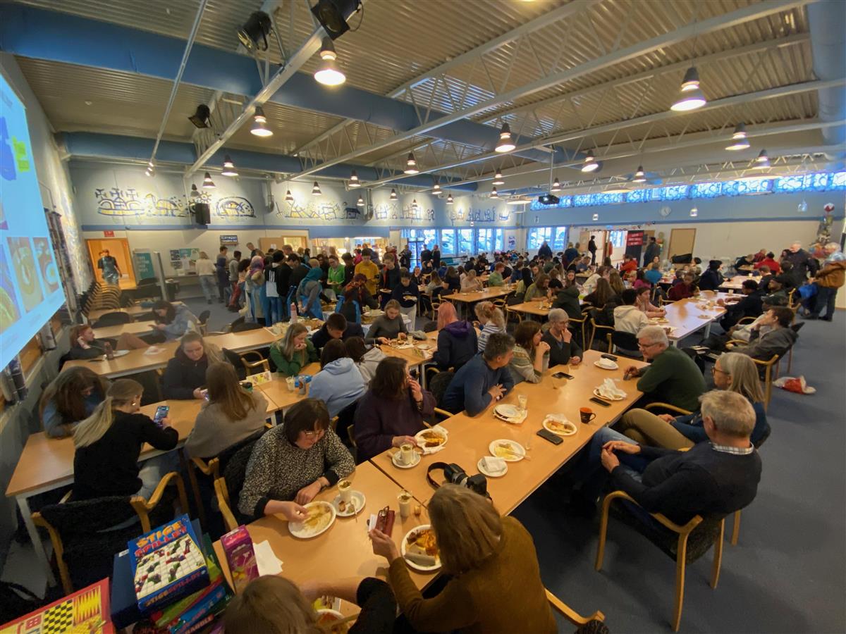 Mange mennesker sitter og spiser i et stor rom/kantine. Fotografi. - Klikk for stort bilde
