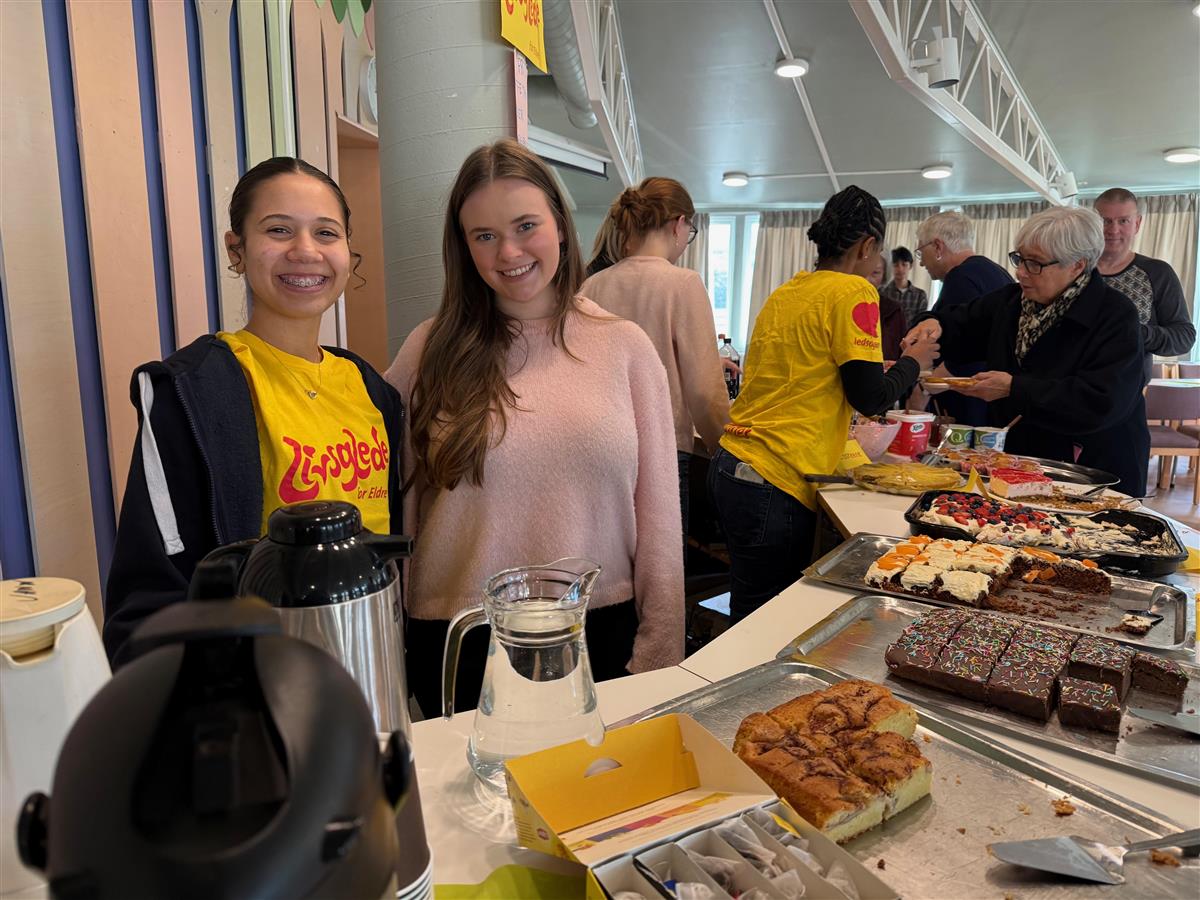 To smilende jenter står bak en kafédisk, flere personer står i bakgrunnen.  - Klikk for stort bilde