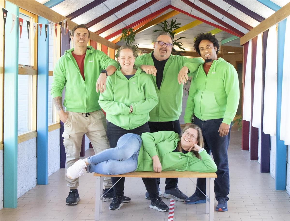 En grønnkledd person ligger på en benk, mens fire andre grønnkledte står bak i en fargerik korridor.  - Klikk for stort bilde